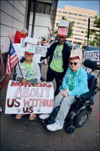Two ADAPTers in wheelchairs and one standing ADAPTer, wearing house shaped cardboard hats, holding a “Nothing About Us Without Us” sign